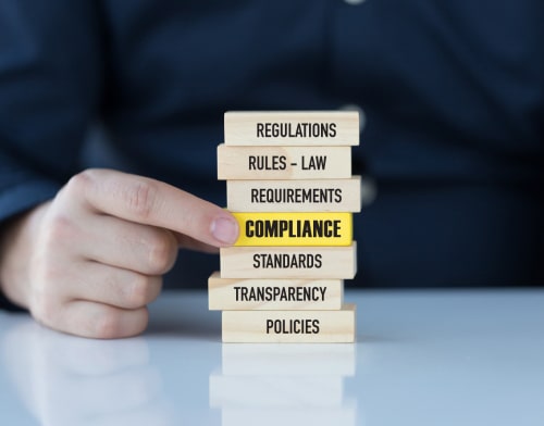 A hand stacking wooden blocks, the middle one being a yellow one with compliance written on it