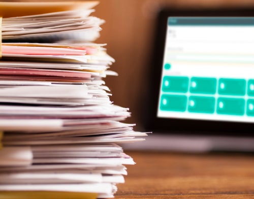 A stack of documents in the foreground, with a laptop in the background