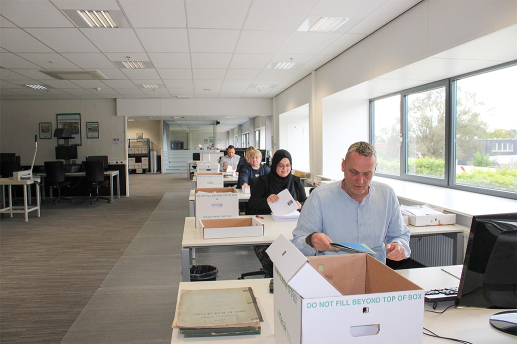 A focused team organizes documents in boxes in a bright office. Natural light streams in from large windows. Efficient and spacious workspace.